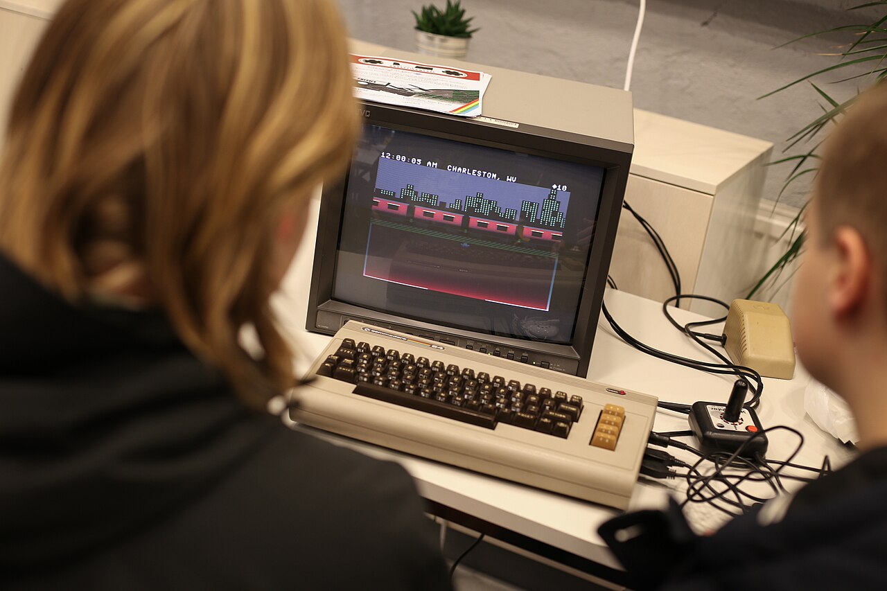 Dwie osoby grają na Commodore 64 z joystickiem przed małym monitorem