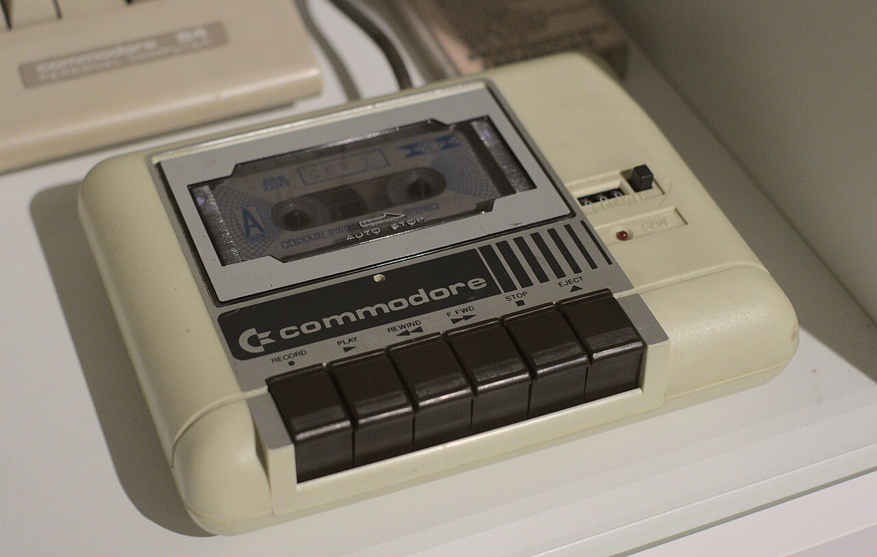 Kremowy magnetofon Commodore Datasette z kasetą w kieszeni i rzędem klawiszy, eksponat muzealny